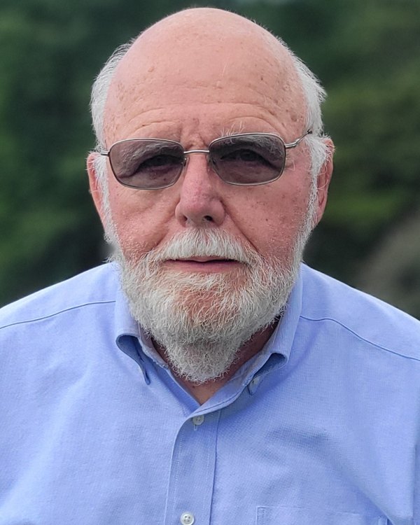 Mark McKeon, with a white beard and sunglasses, in a light blue shirt outdoors.