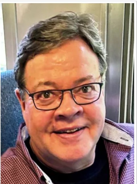 Preston Bristow, smiling in glasses and a plaid shirt, seated aboard an Amtrak coach.
