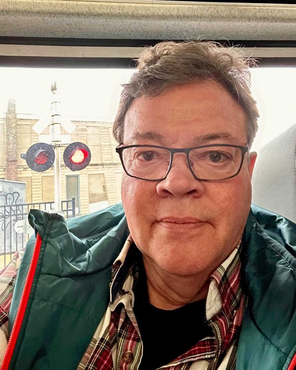 Preston Bristow, in glasses and a plaid shirt, seated aboard a train with a grade-crossing signal outside the window.