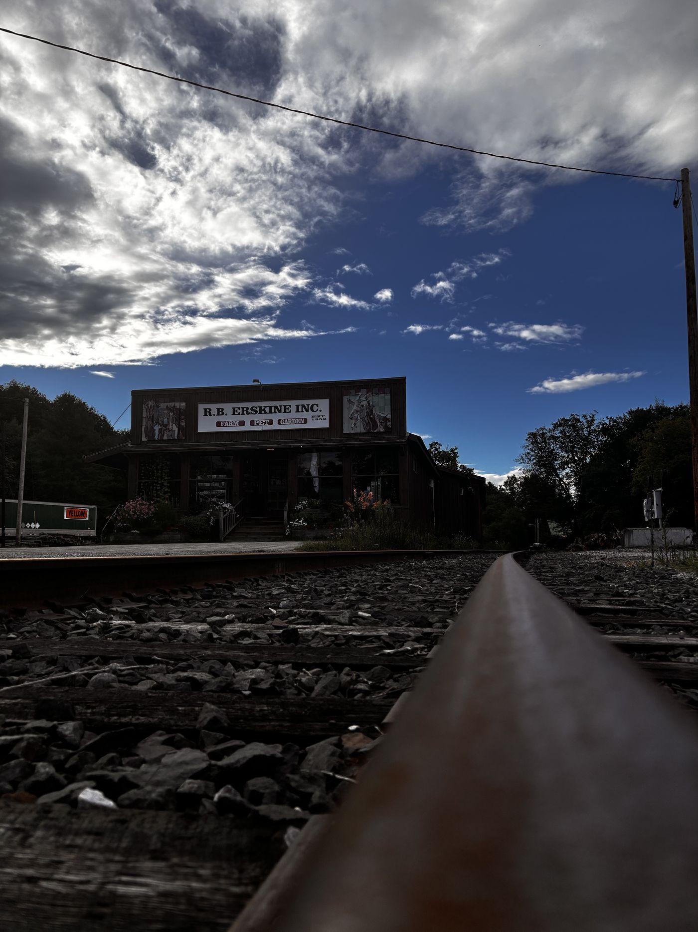 A rail seen from track level, running toward the R.B. Erskine farm and garden store across the road under a dark blue sky.