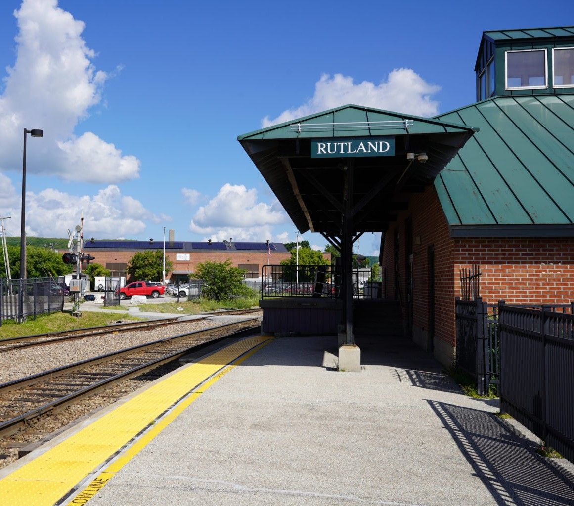 The Rutland Amtrak station: a brick depot with a green metal roof and RUTLAND sign over the platform canopy, tracks and a yellow tactile platform edge in the foreground.