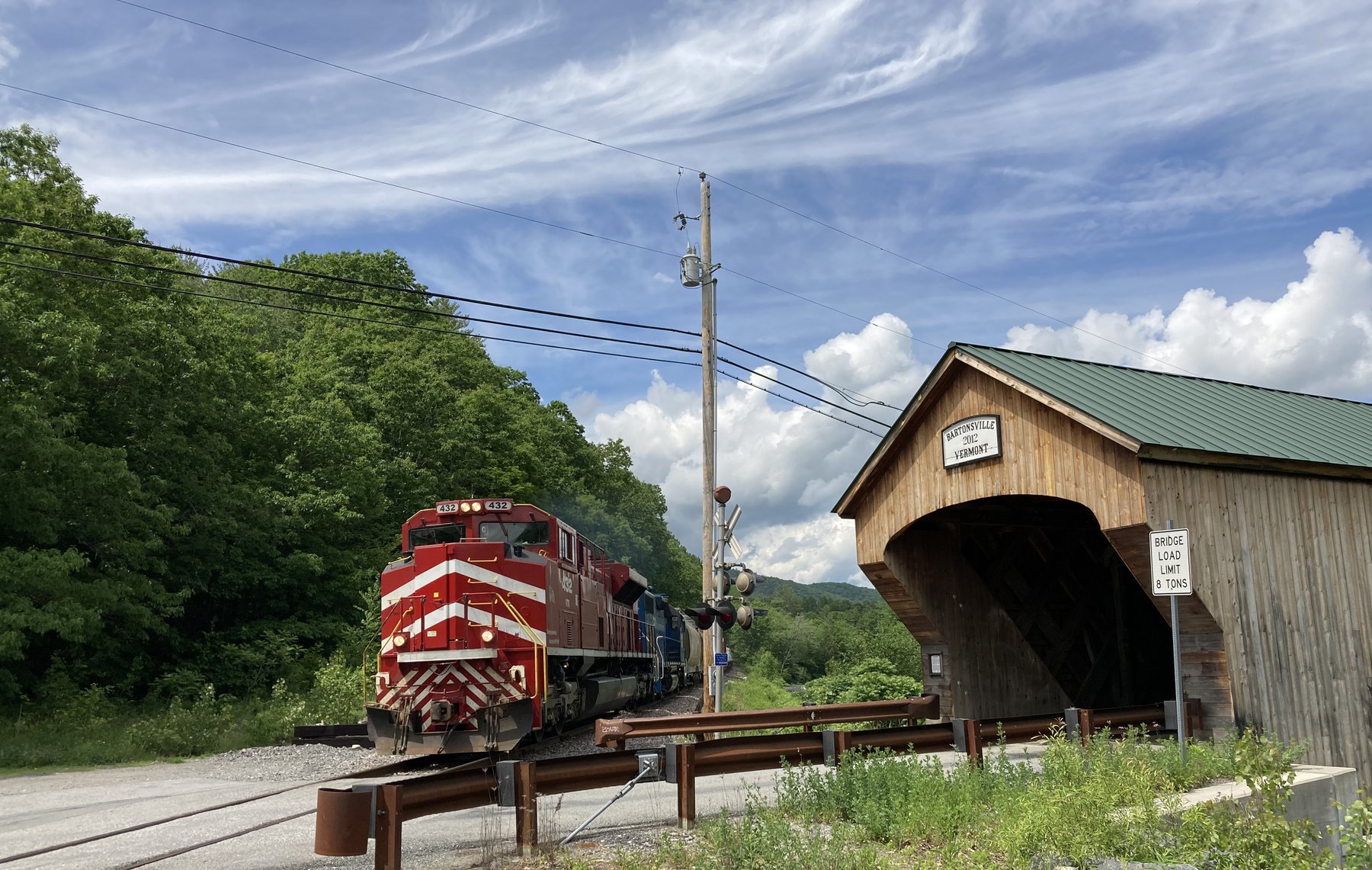A red Vermont Rail System freight locomotive leads a train past the Bartonsville covered bridge on a summer day, the crossing signal between them.
