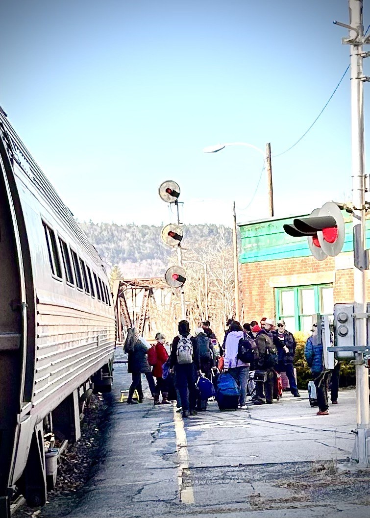 Passengers with luggage gather on the Bellows Falls platform beside a stainless-steel Amtrak coach, with the crossing signals, the old station house and the steel bridge behind them.